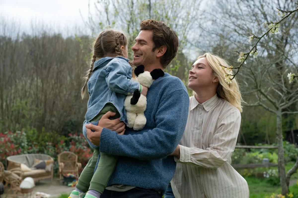 Tobias (Andrew Garfield) and Almut (Florence Pugh) are walking through their backyard, holding their daughter Ella (Grace Delaney).