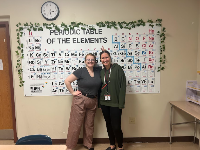 Leah Drennan and Kaitlin Mueller standing in front of the periodic table in their chemistry room.